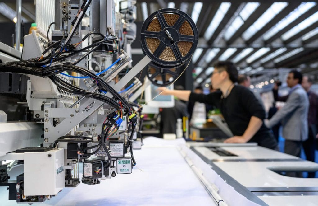 Widok z góry na dużą, przemysłową drukarkę cyfrową podczas pracy. Maszyna zadrukowuje biały materiał kolorowymi grafikami przedstawiającymi sportowców oraz logotypy (m.in. Epson, Summa). W tle widać przestrzeń targową z licznymi stoiskami, przy których pracownicy w niebieskich i zielonych koszulach obsługują urządzenia i rozmawiają z gośćmi.