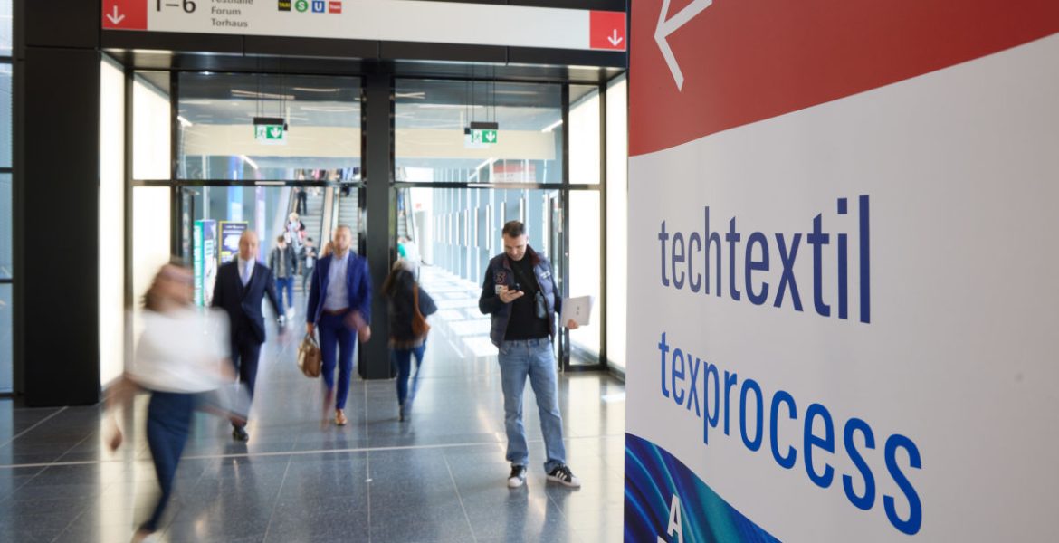 Szeroki kadr na nowoczesne, przeszklone wejście do hali targowej. Na pierwszym planie po prawej stronie znajduje się tablica informacyjna z napisami „techtextil” oraz „texprocess”. W tle widać rozmyte sylwetki odwiedzających poruszających się po lśniącej podłodze oraz znaki kierunkowe prowadzące do sektorów Festhalle i Forum.