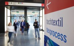Szeroki kadr na nowoczesne, przeszklone wejście do hali targowej. Na pierwszym planie po prawej stronie znajduje się tablica informacyjna z napisami „techtextil” oraz „texprocess”. W tle widać rozmyte sylwetki odwiedzających poruszających się po lśniącej podłodze oraz znaki kierunkowe prowadzące do sektorów Festhalle i Forum.