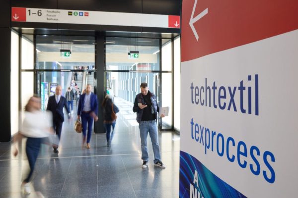 Szeroki kadr na nowoczesne, przeszklone wejście do hali targowej. Na pierwszym planie po prawej stronie znajduje się tablica informacyjna z napisami „techtextil” oraz „texprocess”. W tle widać rozmyte sylwetki odwiedzających poruszających się po lśniącej podłodze oraz znaki kierunkowe prowadzące do sektorów Festhalle i Forum.