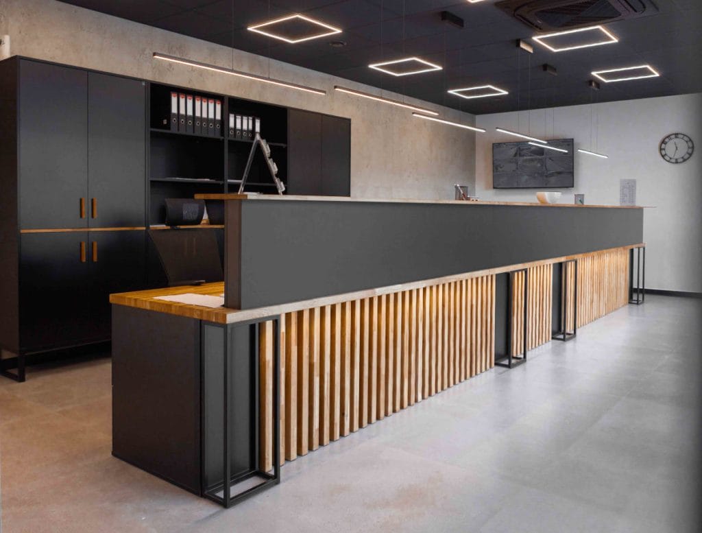 Nowoczesna recepcja biurowa marki Deerhorn z długim ladą wykonaną z pionowych drewnianych lameli i ciemnego blatu. W tle czarne szafy biurowe oraz minimalistyczne, kwadratowe oświetlenie sufitowe LED w loftowym stylu.