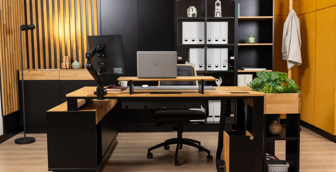 Nowoczesne biuro w stylu loftowym z dużym narożnym biurkiem wykonanym z jasnego drewna i czarnego metalu. Na blacie znajduje się laptop, monitor na regulowanym ramieniu oraz nadstawka. W tle wysoki regał z segregatorami i dekoracyjna ściana z drewnianych lameli.
