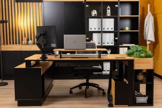 Nowoczesne biuro w stylu loftowym z dużym narożnym biurkiem wykonanym z jasnego drewna i czarnego metalu. Na blacie znajduje się laptop, monitor na regulowanym ramieniu oraz nadstawka. W tle wysoki regał z segregatorami i dekoracyjna ściana z drewnianych lameli.