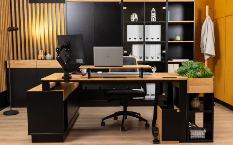 Nowoczesne biuro w stylu loftowym z dużym narożnym biurkiem wykonanym z jasnego drewna i czarnego metalu. Na blacie znajduje się laptop, monitor na regulowanym ramieniu oraz nadstawka. W tle wysoki regał z segregatorami i dekoracyjna ściana z drewnianych lameli.