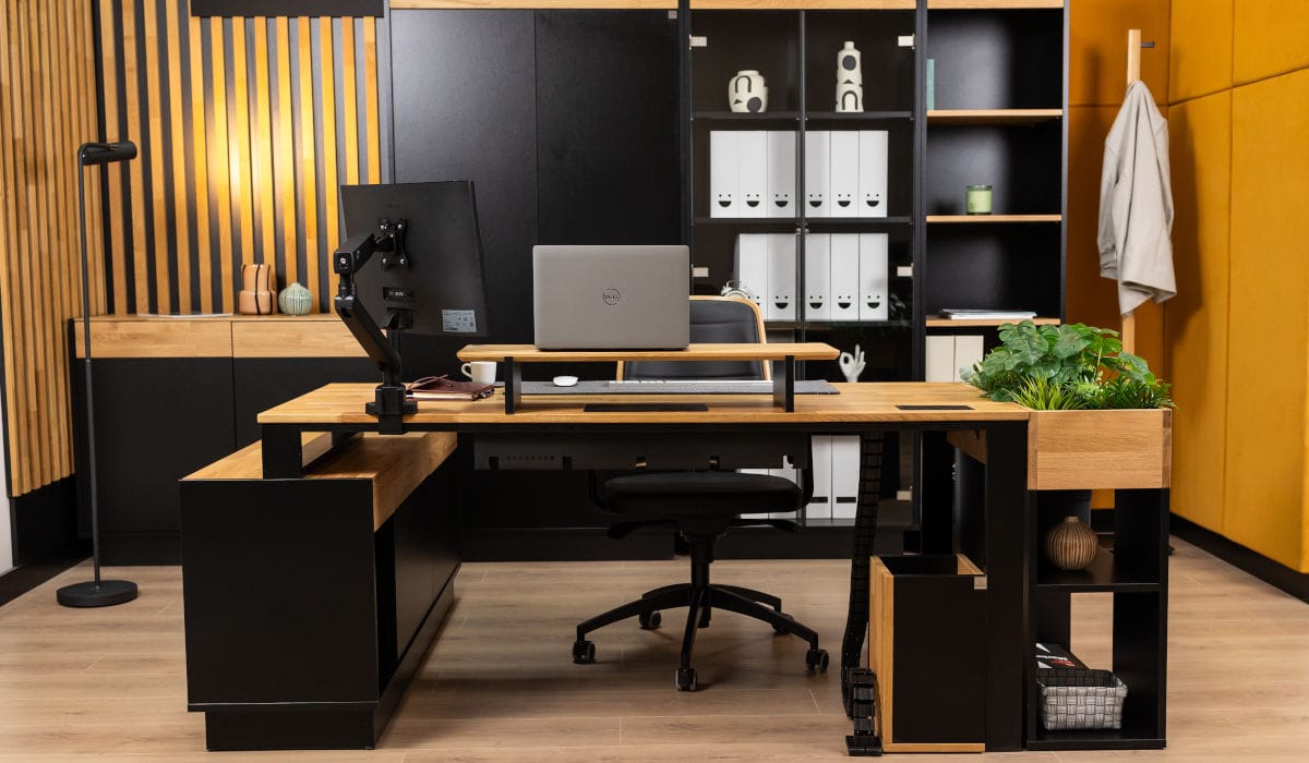 Nowoczesne biuro w stylu loftowym z dużym narożnym biurkiem wykonanym z jasnego drewna i czarnego metalu. Na blacie znajduje się laptop, monitor na regulowanym ramieniu oraz nadstawka. W tle wysoki regał z segregatorami i dekoracyjna ściana z drewnianych lameli.