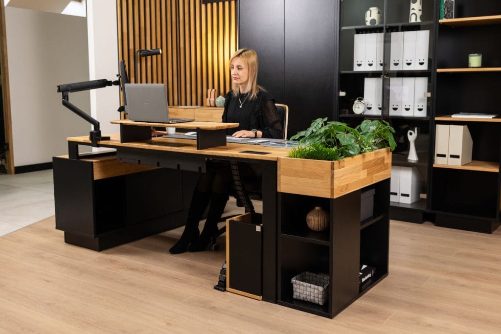 Kobieta o blond włosach siedząca przy nowoczesnym biurku loftowym, pracująca na laptopie. Biurko posiada zintegrowaną donicę z zielonymi roślinami oraz boczne półki. Wnętrze jest utrzymane w kolorystyce dębu, czerni i ciepłej żółci.