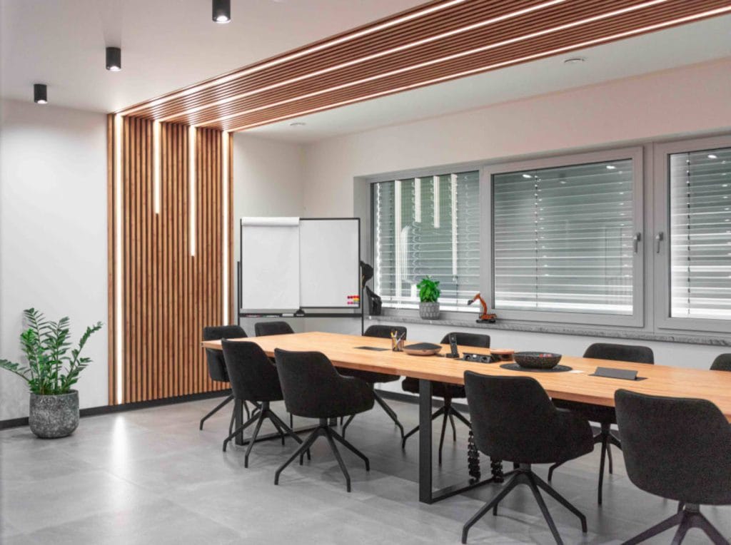 Nowoczesna sala konferencyjna z dużym stołem z litego drewna na czarnych, metalowych nogach. Ściana i fragment sufitu ozdobione pionowymi drewnianymi lamelami z wbudowanym oświetleniem liniowym. W pomieszczeniu znajdują się tapicerowane czarne krzesła obrotowe.