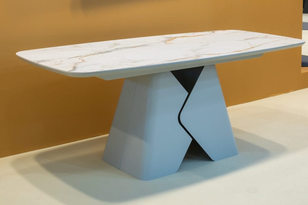 Nowoczesny stół Prisma z blatem o wzorze białego marmuru z szarymi i złotymi żyłkami. Podstawa stołu ma unikalną, masywną formę geometryczną w kolorze jasnoszarym.