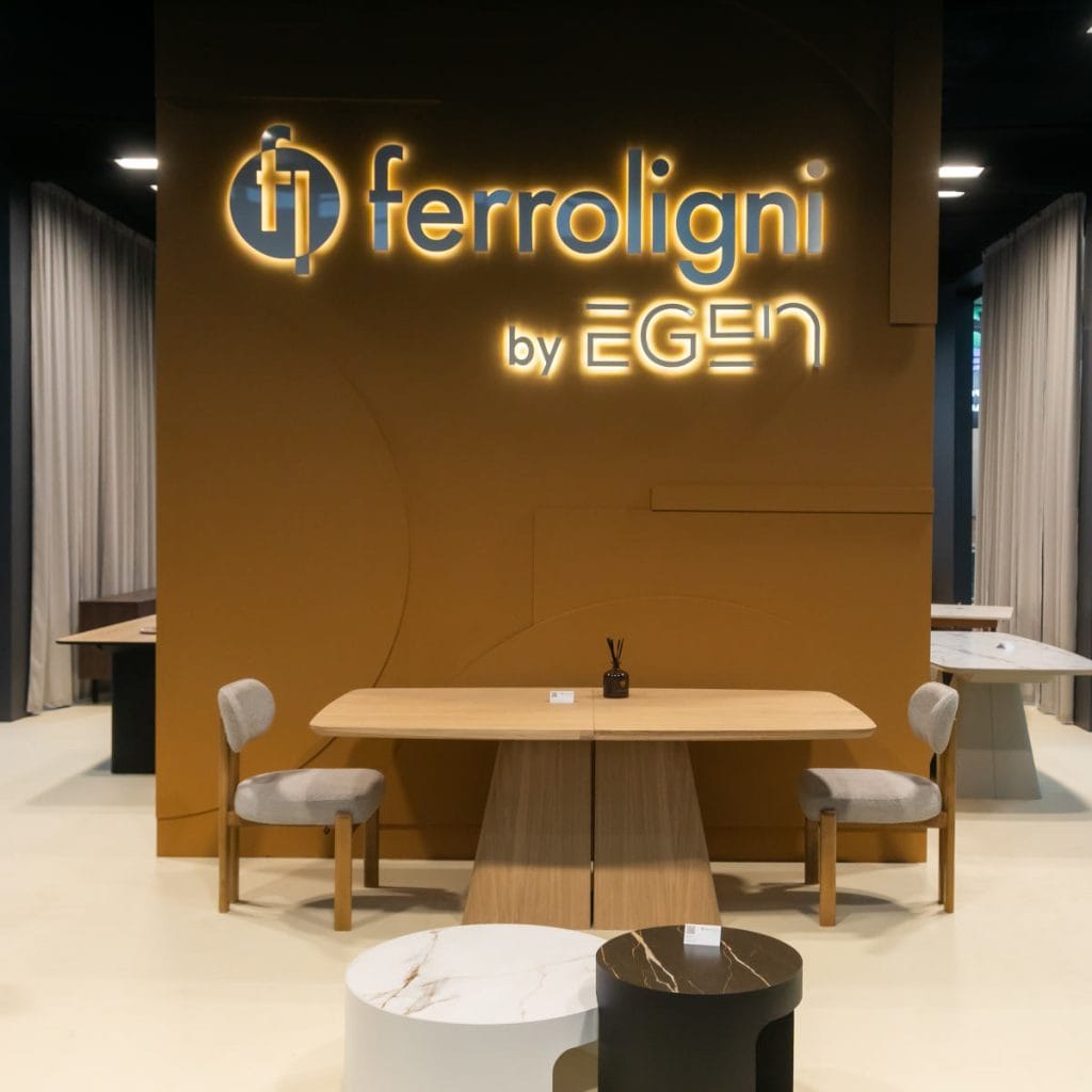 Ekspozycja mebli Ferroligni: drewniany stół z krzesłami na tle pomarańczowej ściany z logo marki. Na pierwszym planie widoczne niskie stoliki kawowe z marmurowymi blatami.