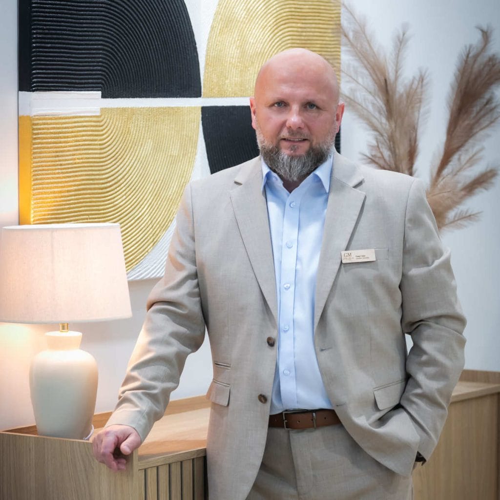 Brodaty mężczyzna w beżowym garniturze i jasnoniebieskiej koszuli stoi na tle abstrakcyjnego obrazu w odcieniach złota, czerni i bieli oraz suszonej trawy pampasowej. Jedną rękę trzyma na szafce z jasnego drewna, a drugą w kieszeni. Na jego marynarce przypięty jest identyfikator.