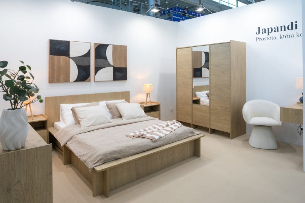 toisko targowe prezentujące sypialnię w stylu Japandi z łóżkiem z jasnego drewna, dwiema szafkami nocnymi z lampami, szafą z lustrem, białą pufą i toaletką. Nad łóżkiem wiszą dwa abstrakcyjne obrazy, a obok niego stoi biały wazon z gałązką eukaliptusa.