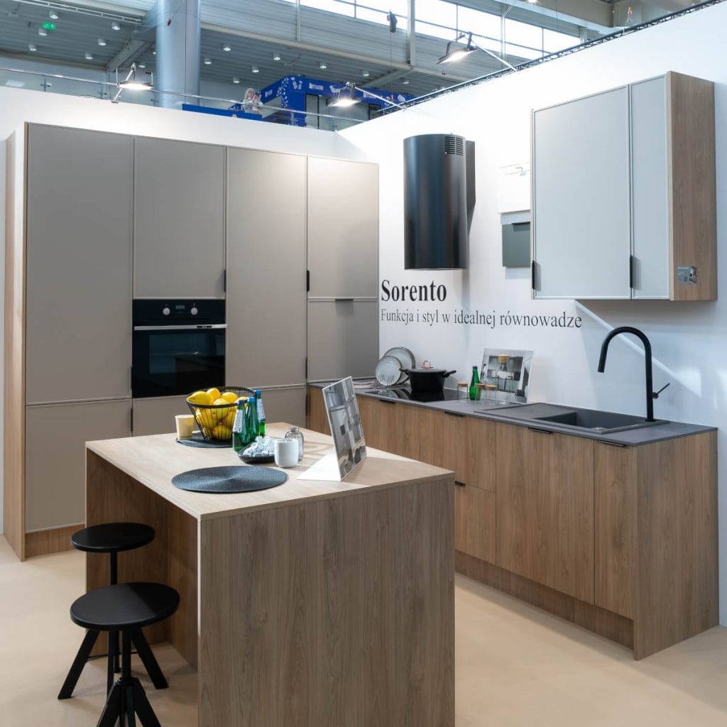 Aranżacja nowoczesnej kuchni z szafkami w kolorze jasnego drewna i matowej szarości. Wyposażona jest w czarny piekarnik, płytę grzewczą, okrągły okap, zlew z czarną baterią oraz wyspę kuchenną z dwoma hokerami i koszem owoców. Na ścianie widać fragment napisu "Sorento".