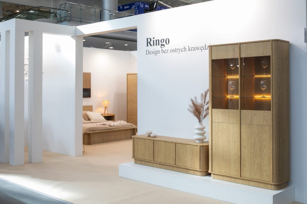 Stoisko targowe prezentujące drewnianą witrynę z podświetlanymi półkami, na których stoją kieliszki, oraz pasującą do niej niską komodę. Na komodzie stoi biały karbowany wazon z trawą pampasową. W tle widać fragment sypialni.