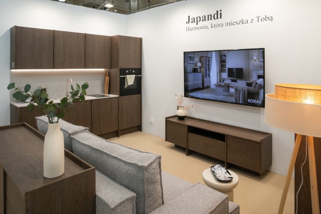 Aranżacja salonu i aneksu kuchennego w ciemnym drewnie. Widoczna jest szara, modułowa sofa, niska szafka RTV z telewizorem, fragmenty ciemnych mebli kuchennych oraz wysoka lampa stojąca z abażurem. Na ścianie wisi napis "Japandi".