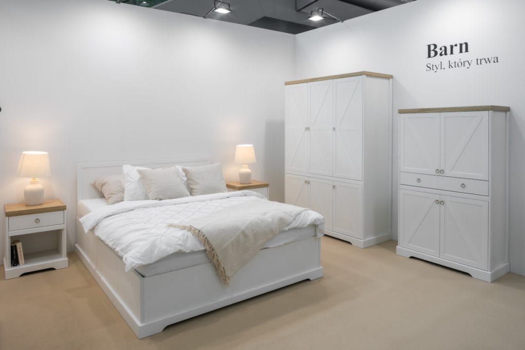 Aranżacja sypialni Barn z białym łóżkiem, dwiema szafkami nocnymi z lampami, wysoką białą szafą i komodą w stylu rustykalnym. Meble mają charakterystyczne fronty z krzyżakami. Na białej ścianie widać czarny napis "Barn".