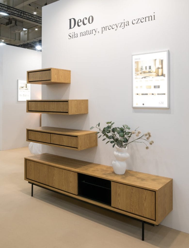 Stoisko targowe z nowoczesną niską szafką RTV z drewna dębowego na czarnych metalowych nogach. Nad szafką wiszą trzy dębowe półki szufladowe. Na szafce stoi biały wazon z gałązką eukaliptusa, a nad nią wiszą dwa podświetlane plakaty i napis "Deco".