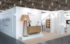 Szerokie ujęcie stoiska targowego firmy GAŁA MEBLE. Na białej ścianie widoczne jest logo i adres strony internetowej, a obok stojąca lampa z dużym abażurem, dębowa komoda i witryna. W tle widać dalszą część stoiska z białymi filarami.