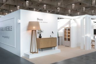 Szerokie ujęcie stoiska targowego firmy GAŁA MEBLE. Na białej ścianie widoczne jest logo i adres strony internetowej, a obok stojąca lampa z dużym abażurem, dębowa komoda i witryna. W tle widać dalszą część stoiska z białymi filarami.