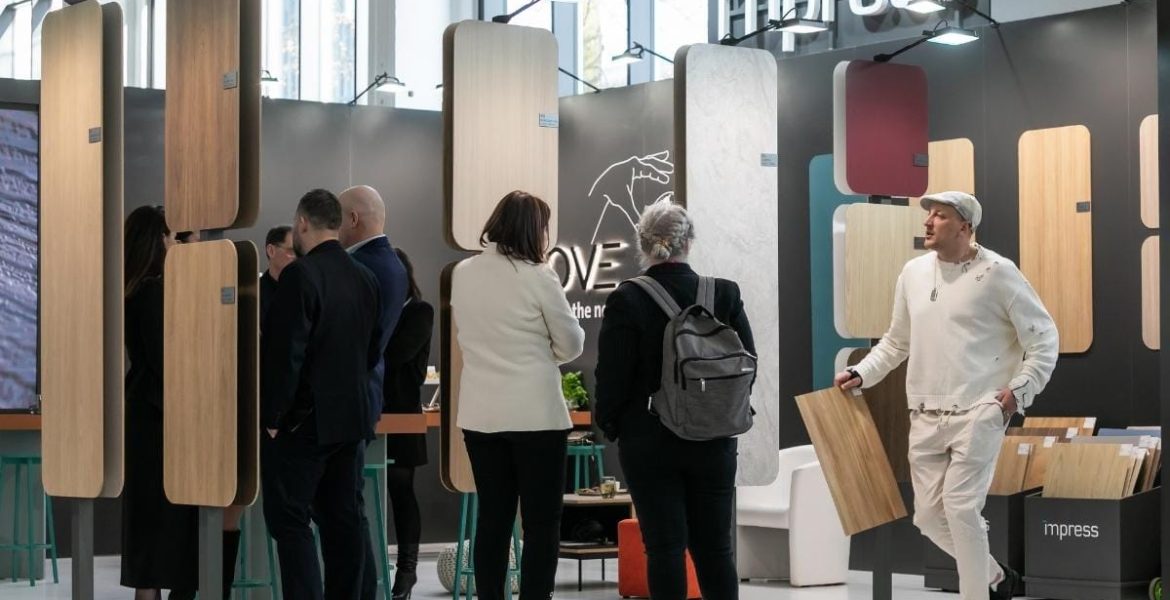 Perspektywa na ścianę z wzornikami na stoisku Impress Decor. Na pierwszym planie dekor Goldstone Oak (32407). W tle widoczni specjaliści branżowi podczas rozmów, co obrazuje intensywne targi meblowe i aktywne nawiązywanie relacji biznesowych.