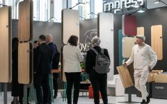 Perspektywa na ścianę z wzornikami na stoisku Impress Decor. Na pierwszym planie dekor Goldstone Oak (32407). W tle widoczni specjaliści branżowi podczas rozmów, co obrazuje intensywne targi meblowe i aktywne nawiązywanie relacji biznesowych.