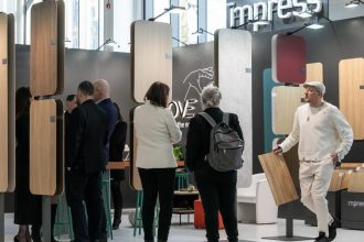 Perspektywa na ścianę z wzornikami na stoisku Impress Decor. Na pierwszym planie dekor Goldstone Oak (32407). W tle widoczni specjaliści branżowi podczas rozmów, co obrazuje intensywne targi meblowe i aktywne nawiązywanie relacji biznesowych.