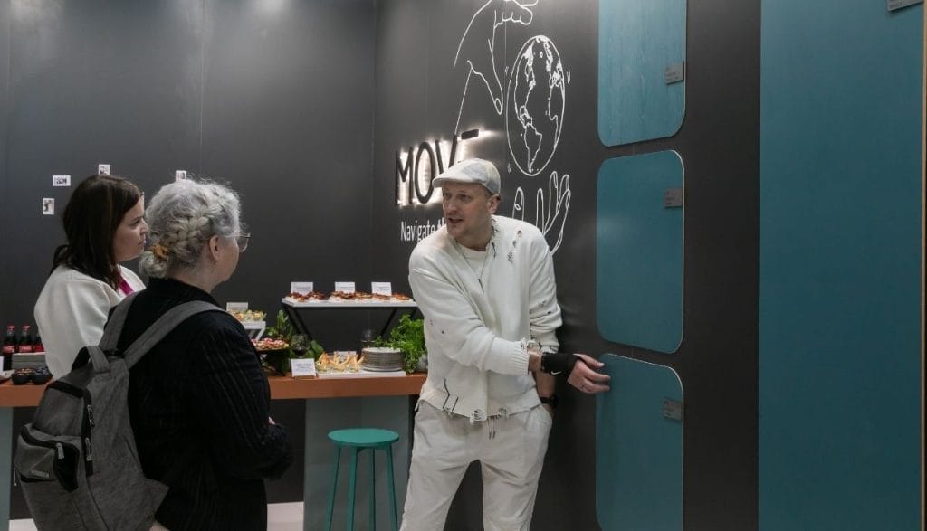 Rozmowa handlowa na stoisku Impress Decor. Przedstawiciel firmy prezentuje klientkom dekory w kolorze petrol na tle logotypu MOVE. W tle widoczna strefa cateringowa, co podkreśla partnerskie relacje biznesowe i komfortowe warunki współpracy podczas targów meblowych.