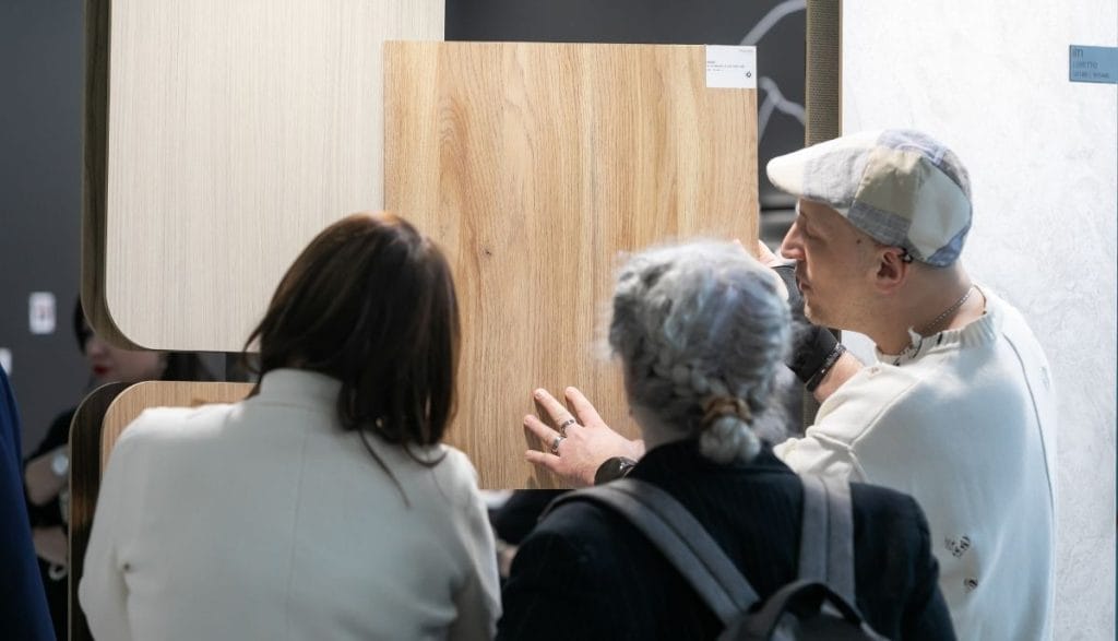 Prezentacja jakości dekoru dębowego na stoisku Impress Decor. Klientki oceniają strukturę i usłojenie dużej płyty meblowej trzymanej przez doradcę. Bezpośredni kontakt z produktem to kluczowy element budowania zaufania i relacji biznesowych na targach meblowych.