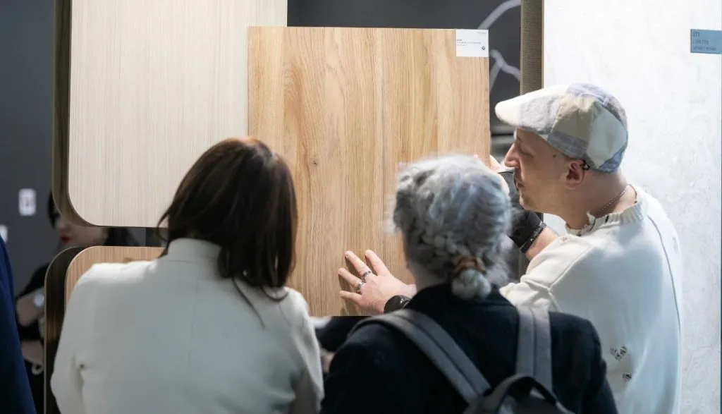 Prezentacja jakości dekoru dębowego na stoisku Impress Decor. Klientki oceniają strukturę i usłojenie dużej płyty meblowej trzymanej przez doradcę. Bezpośredni kontakt z produktem to kluczowy element budowania zaufania i relacji biznesowych na targach meblowych.