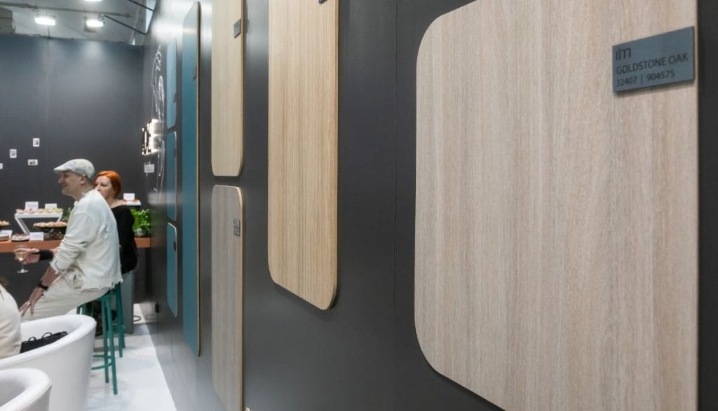 Szerokie ujęcie stoiska Impress Decor na targach Meble Polska 2026. Eksperci firmy prezentują nową kolekcję dekorów dębowych grupie profesjonalistów. Zdjęcie oddaje dynamiczne targi meblowe i profesjonalne relacje biznesowe w nowoczesnej przestrzeni wystawienniczej.