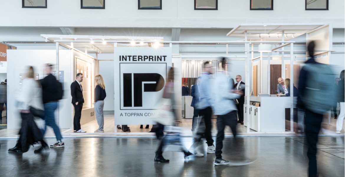 Szerokie ujęcie stoiska targowego firmy Interprint. Na pierwszym planie widać rozmazane sylwetki przechodzących ludzi, co nadaje zdjęciu dynamiki. W centrum znajduje się duże białe logo "IP" z napisem "INTERPRINT - A TOPPAN COMPANY". Konstrukcja stoiska jest jasna i minimalistyczna.