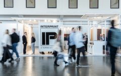 Szerokie ujęcie stoiska targowego firmy Interprint. Na pierwszym planie widać rozmazane sylwetki przechodzących ludzi, co nadaje zdjęciu dynamiki. W centrum znajduje się duże białe logo "IP" z napisem "INTERPRINT - A TOPPAN COMPANY". Konstrukcja stoiska jest jasna i minimalistyczna.