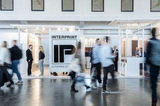 Szerokie ujęcie stoiska targowego firmy Interprint. Na pierwszym planie widać rozmazane sylwetki przechodzących ludzi, co nadaje zdjęciu dynamiki. W centrum znajduje się duże białe logo "IP" z napisem "INTERPRINT - A TOPPAN COMPANY". Konstrukcja stoiska jest jasna i minimalistyczna.