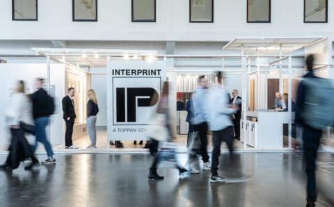 Szerokie ujęcie stoiska targowego firmy Interprint. Na pierwszym planie widać rozmazane sylwetki przechodzących ludzi, co nadaje zdjęciu dynamiki. W centrum znajduje się duże białe logo "IP" z napisem "INTERPRINT - A TOPPAN COMPANY". Konstrukcja stoiska jest jasna i minimalistyczna.