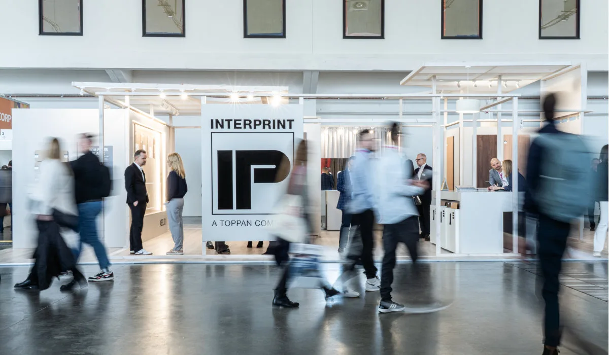 Szerokie ujęcie stoiska targowego firmy Interprint. Na pierwszym planie widać rozmazane sylwetki przechodzących ludzi, co nadaje zdjęciu dynamiki. W centrum znajduje się duże białe logo "IP" z napisem "INTERPRINT - A TOPPAN COMPANY". Konstrukcja stoiska jest jasna i minimalistyczna.