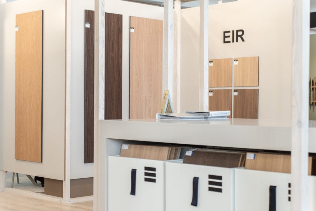 Zbliżenie na ekspozycję próbek dekorów meblowych. Pionowe panele o różnych odcieniach drewna zamontowane na jasnych ściankach. Na dole widoczne białe szafki z uchwytami w formie czarnych pasków, wypełnione wzornikami. Nad jedną z sekcji widnieje napis "EIR".