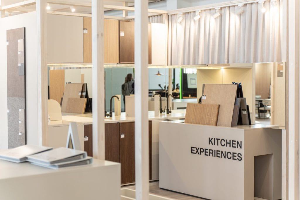 Widok na sekcję stoiska o nazwie "KITCHEN EXPERIENCES". Aranżacja przypomina nowoczesną kuchnię z ciemnobrązowymi szafkami i jasnym blatem. Na blacie wyeksponowano próbki blatów i frontów oraz czarną baterię kuchenną. Całość oświetlona punktowymi reflektorami.