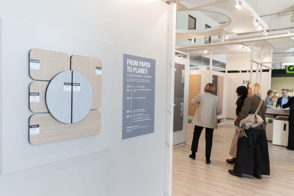 Tablica informacyjna pod tytułem "FROM PAPER TO PLANET: A SUSTAINABLE JOURNEY" prezentująca oś czasu działań proekologicznych firmy Interprint. Obok tablicy znajduje się dekoracja z próbek materiałów w kształcie koła i zaokrąglonych prostokątów. W tle widać odwiedzających stoisko.