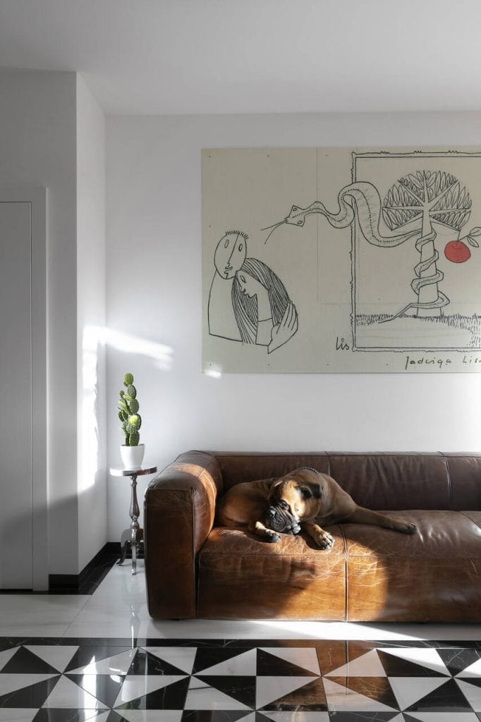 Brązowa skórzana sofa, na której odpoczywa suczka – ukochana towarzyszka domowników – przyciąga uwagę elegancją i komfortem. Na ścianie wisi wyjątkowa grafika matki właścicieli, nadając wnętrzu osobisty i artystyczny charakter. Połączenie klasyki i subtelnych detali tworzy przestrzeń pełną ciepła i indywidualności.