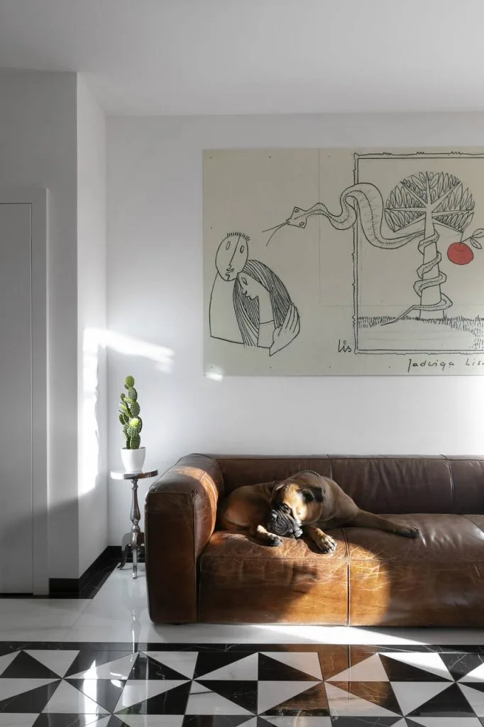 Brązowa skórzana sofa, na której odpoczywa suczka – ukochana towarzyszka domowników – przyciąga uwagę elegancją i komfortem. Na ścianie wisi wyjątkowa grafika matki właścicieli, nadając wnętrzu osobisty i artystyczny charakter. Połączenie klasyki i subtelnych detali tworzy przestrzeń pełną ciepła i indywidualności.