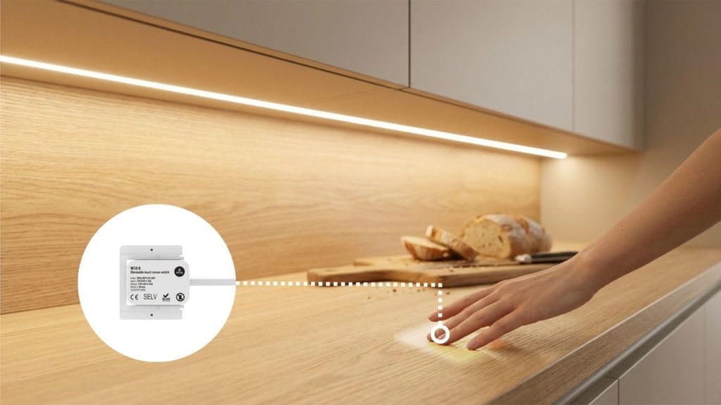 Zbliżenie na dłoń dotykającą drewnianego blatu w jasnej kuchni, tuż pod listwą LED. Obok w białym kole widoczny jest schemat techniczny urządzenia sterującego oświetleniem, połączony linią przerywaną z miejscem dotyku. W tle widać deskę do krojenia z chlebem.