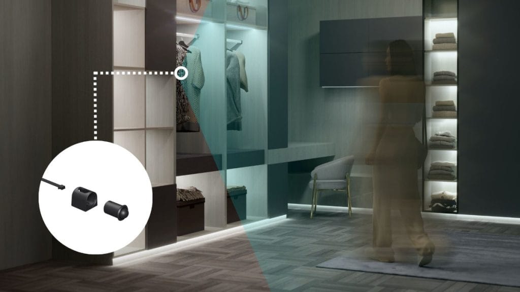 Wnętrze nowoczesnej garderoby z szafami podświetlonymi chłodnym, niebieskim światłem LED. Rozmazana sylwetka kobiety idzie wzdłuż szaf. Po lewej stronie w białym kole pokazane są elementy złącza elektrycznego, połączone linią ze strefą oświetlenia.