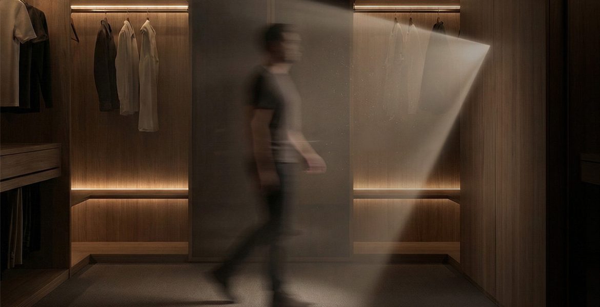 Rozmazana sylwetka mężczyzny przechodzącego przez luksusową, ciemną garderobę wykończoną drewnem. Ciepłe światło z wbudowanych listew LED oświetla wiszące ubrania i podłogę, a snop światła pada również z prawej strony.