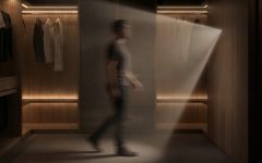 Rozmazana sylwetka mężczyzny przechodzącego przez luksusową, ciemną garderobę wykończoną drewnem. Ciepłe światło z wbudowanych listew LED oświetla wiszące ubrania i podłogę, a snop światła pada również z prawej strony.
