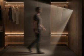 Rozmazana sylwetka mężczyzny przechodzącego przez luksusową, ciemną garderobę wykończoną drewnem. Ciepłe światło z wbudowanych listew LED oświetla wiszące ubrania i podłogę, a snop światła pada również z prawej strony.