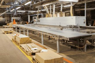 Park maszynowy w nowoczesnym zakładzie obróbki drewna. Widoczna linia produkcyjna do prefabrykacji mebli oraz przygotowane komponenty drewniane na paletach wzdłuż ciągu komunikacyjnego.