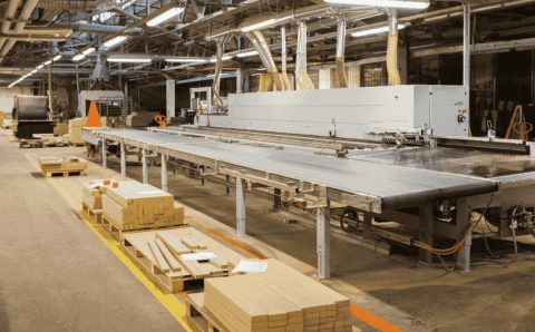 Park maszynowy w nowoczesnym zakładzie obróbki drewna. Widoczna linia produkcyjna do prefabrykacji mebli oraz przygotowane komponenty drewniane na paletach wzdłuż ciągu komunikacyjnego.