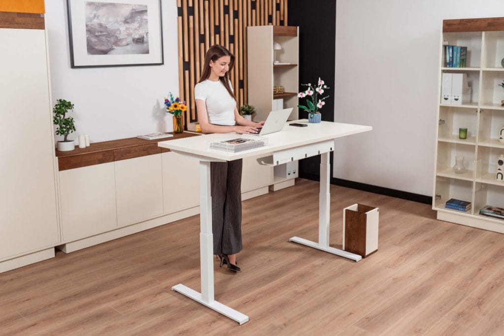 Kobieta pracująca na stojąco przy białym biurku z elektryczną regulacją wysokości. W tle jasne meble biurowe, biały regał z segregatorami oraz dekoracyjne panele ścienne z drewna.