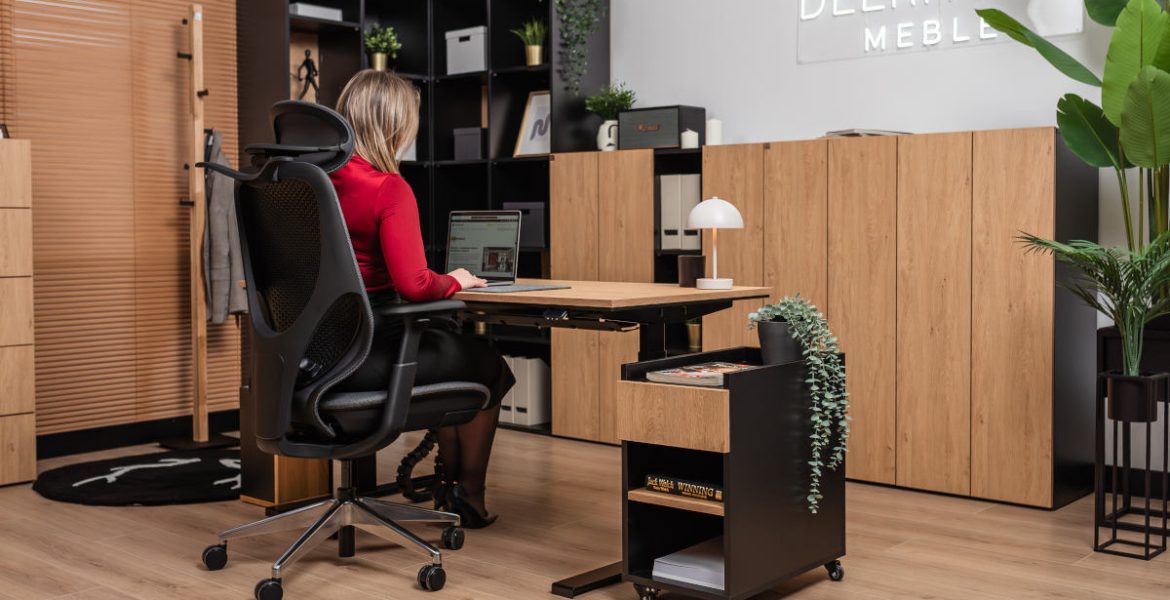 Kobieta siedząca w ergonomicznym fotelu przy dębowym biurku z czarnym stelażem. Obok biurka znajduje się czarny kontener na kółkach z dębowym frontem, a w tle widać nowoczesne regały biurowe i rośliny doniczkowe.