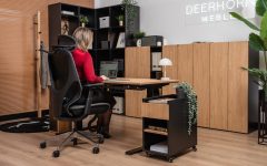 Kobieta siedząca w ergonomicznym fotelu przy dębowym biurku z czarnym stelażem. Obok biurka znajduje się czarny kontener na kółkach z dębowym frontem, a w tle widać nowoczesne regały biurowe i rośliny doniczkowe.