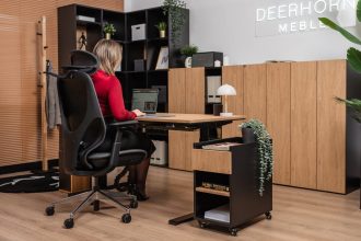 Kobieta siedząca w ergonomicznym fotelu przy dębowym biurku z czarnym stelażem. Obok biurka znajduje się czarny kontener na kółkach z dębowym frontem, a w tle widać nowoczesne regały biurowe i rośliny doniczkowe.